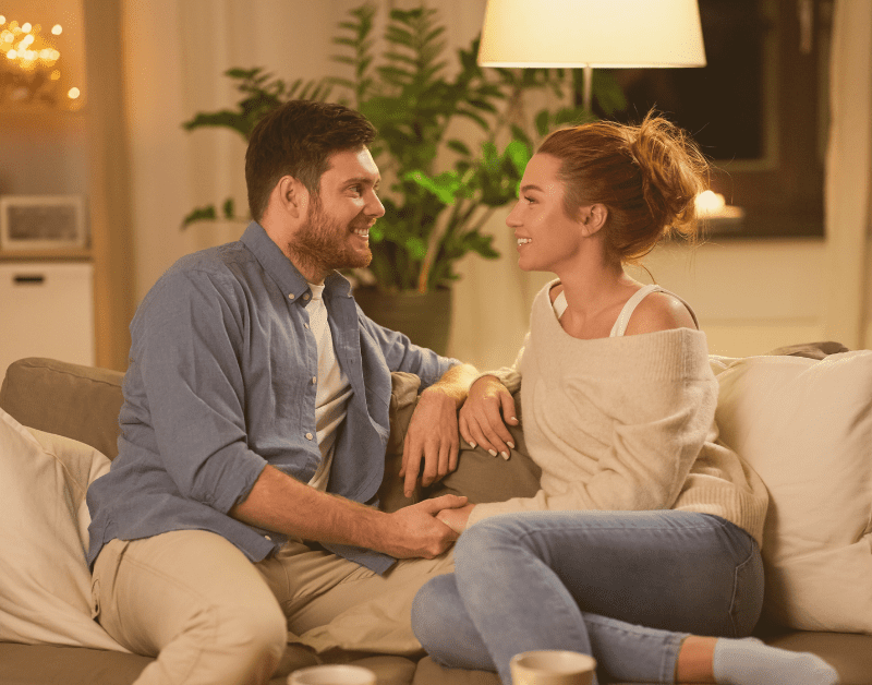 man and woman sitting on couch looking and smiling at each other holding hands